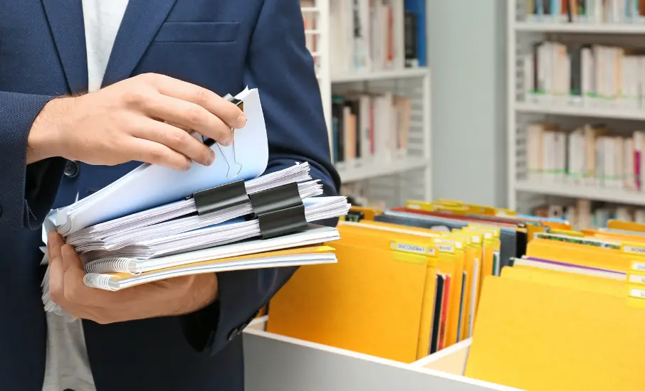 Homme en tenue professionnelle manipulant des dossiers et classeurs dans un bureau, illustrant la gestion documentaire en entreprise.